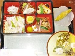 仕切り付きの弁当箱に色とりどりのおかずが詰められている。弁当箱の横にはメロンも添えられている。