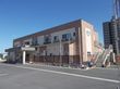屋外階段が設置されたベージュとブラウンの2階建ての建物。玄関には車寄せがあり、建物を囲むようにして花が植えられたプランターが置かれている。