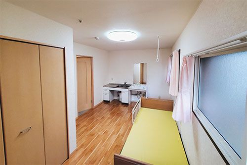ピンクとブラウンの外壁の２階建ての建物の玄関には段差がなく、玄関の屋根ともなっているバルコニーにはホーム名が書かれている。