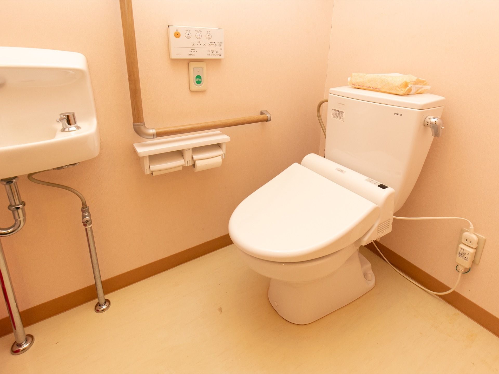トイレには温水洗浄便座が設置されている。壁にはＬ字型の手すりが取り付けられており、横にはコンパクトなタイプの手洗い器が設けられている。