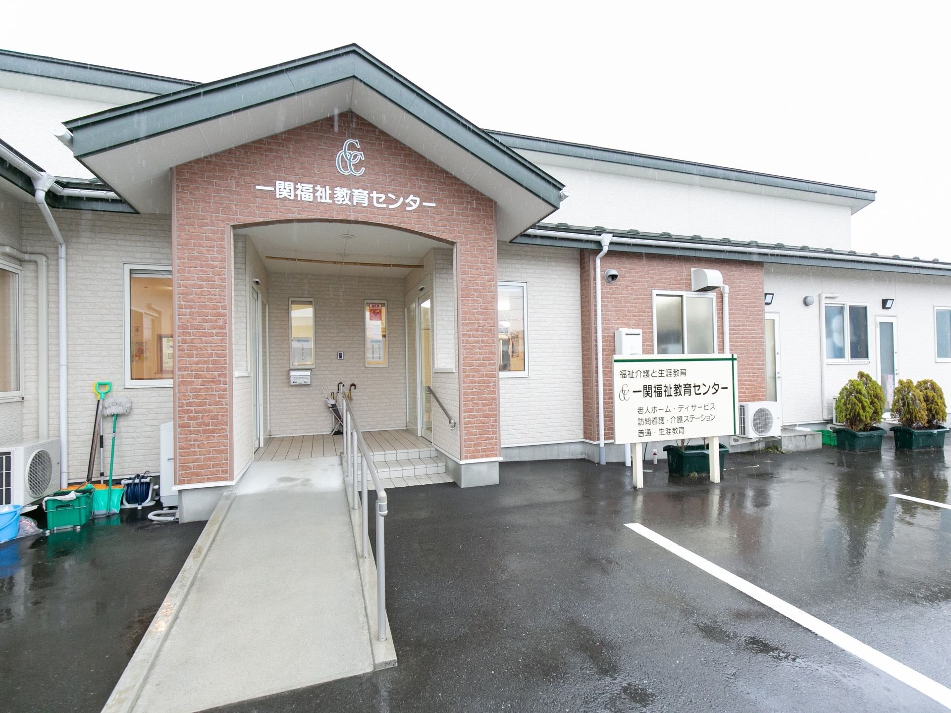 玄関の入口には階段とスロープが設置されている。右側には駐車場が設けられており、施設名の描かれた看板が立てられている。