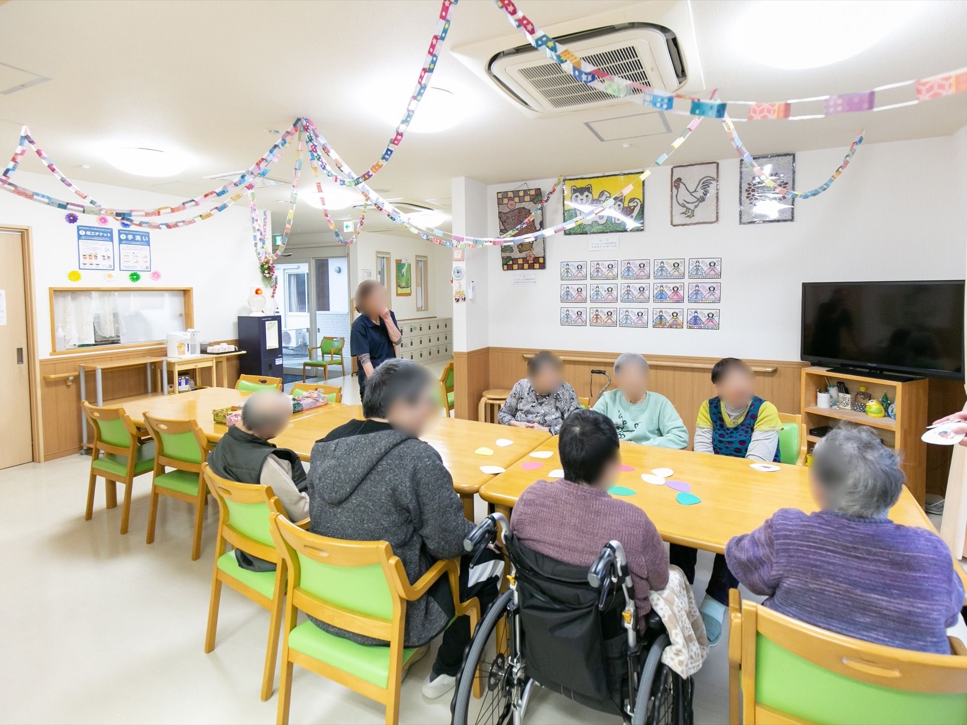 食堂の中央にはダイニングテーブルが並べられており、入居者が座っている。壁には絵などが貼られており、天井には飾り付けがされている。