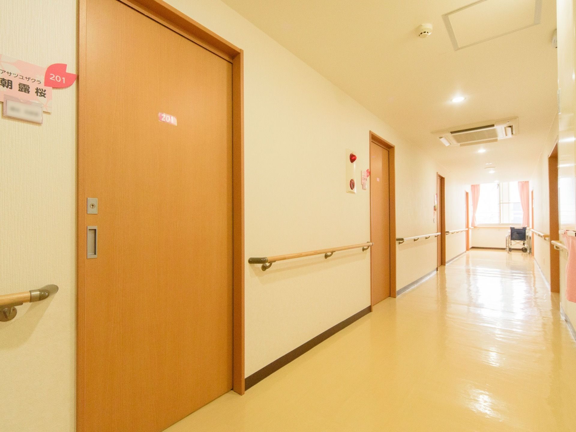木製の引き戸のドアで、左側の壁には部屋番号とともにネームプレートが貼られている。廊下の突き当りには、車椅子がある。
