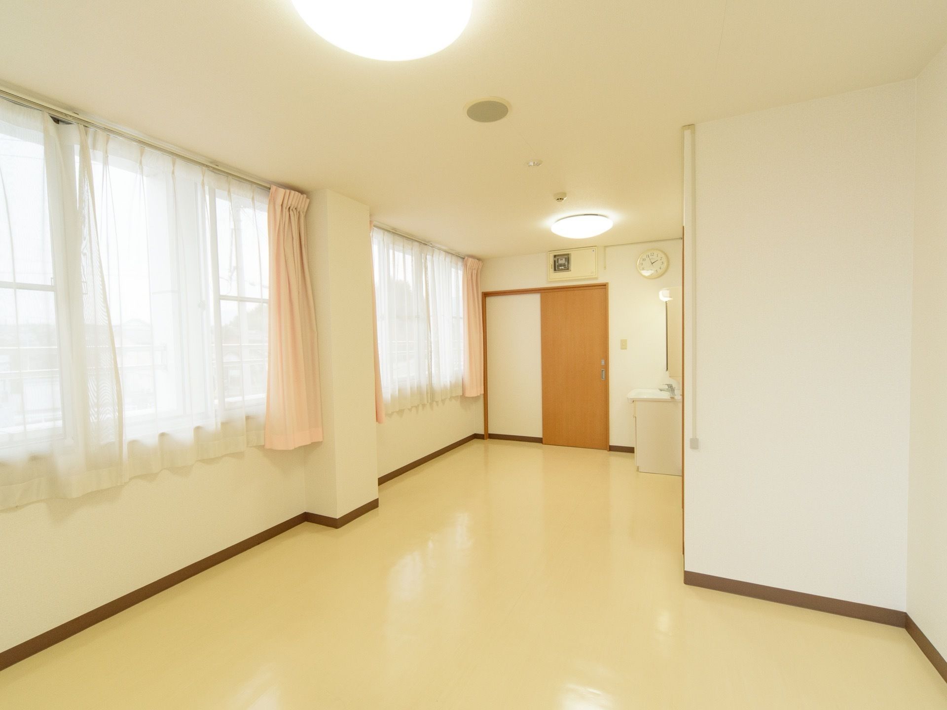 大きな窓が取り付けられており、レースカーテンと薄いピンク色のカーテンが取り付けられている。突き当りには、部屋がある。