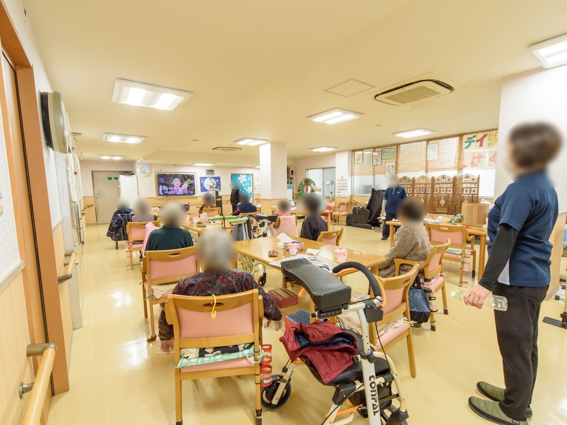 入居者様が椅子に座り、壁に取り付けられたテレビを見ている。奥には、大きなドアの非常口が設置されている。