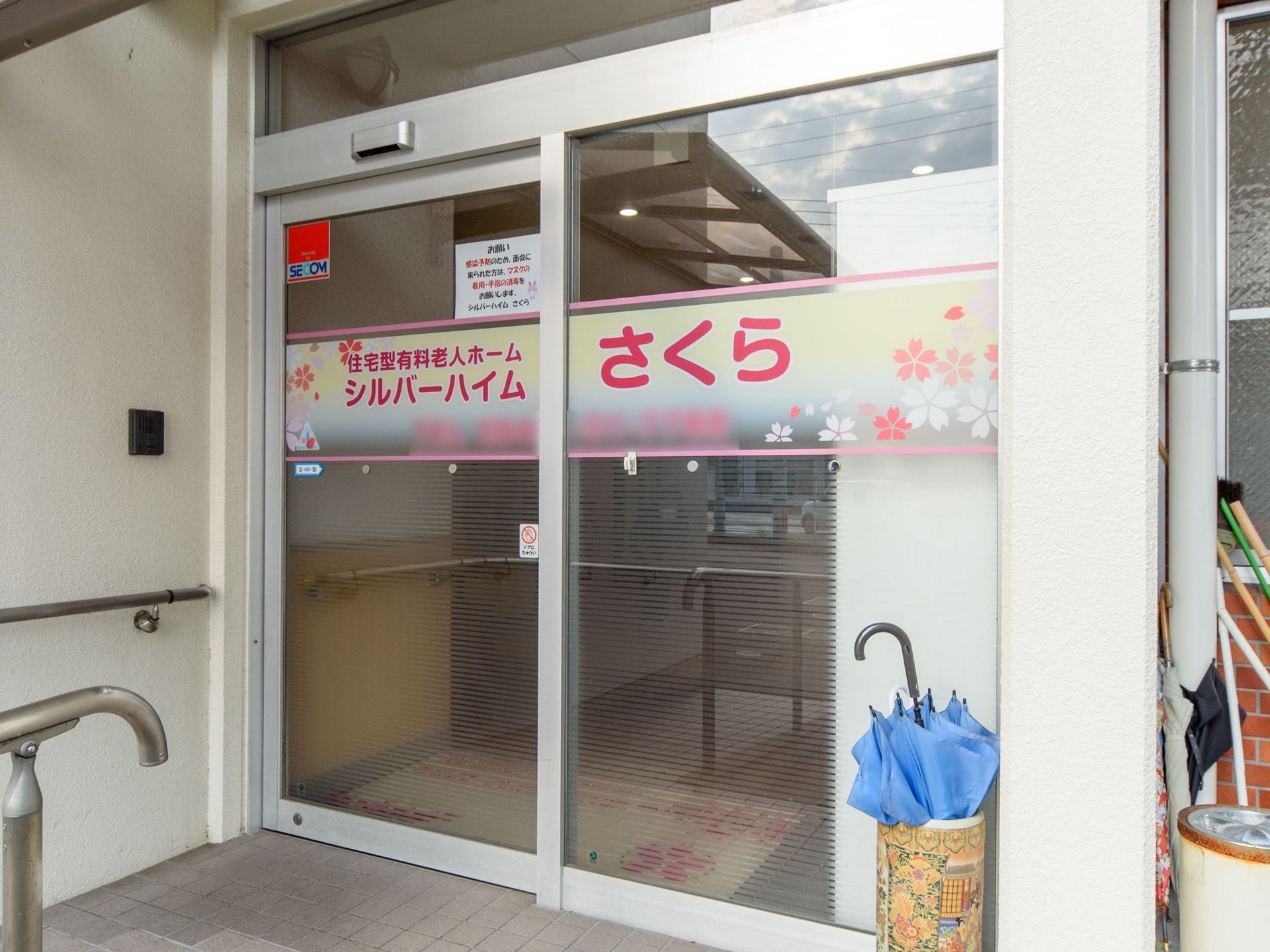 透明なガラスの玄関で、中央には施設名とともに桜のイラストが描かれている。左側の壁には、インターフォンと手すりが付いている。
