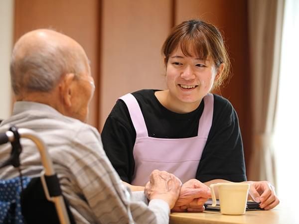 エプロンを付けた若い男女のスタッフに付き添わり、屋外で何かを楽しそうに鑑賞している車いすの高齢の男女。
