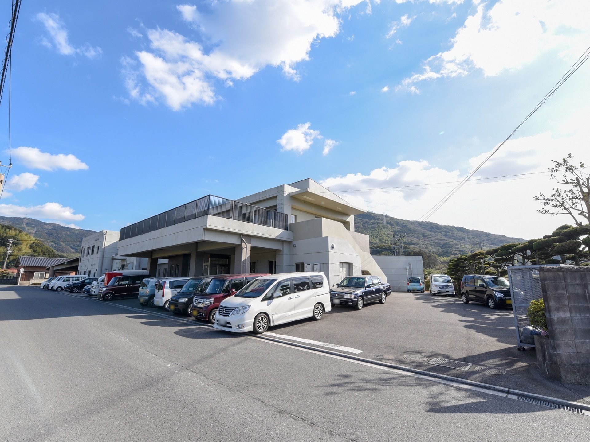 山並みが近くに見える敷地に建つ２階建ての施設である。２階にはとても広いバルコニーがあり、その下は玄関の車寄せにもなっている。