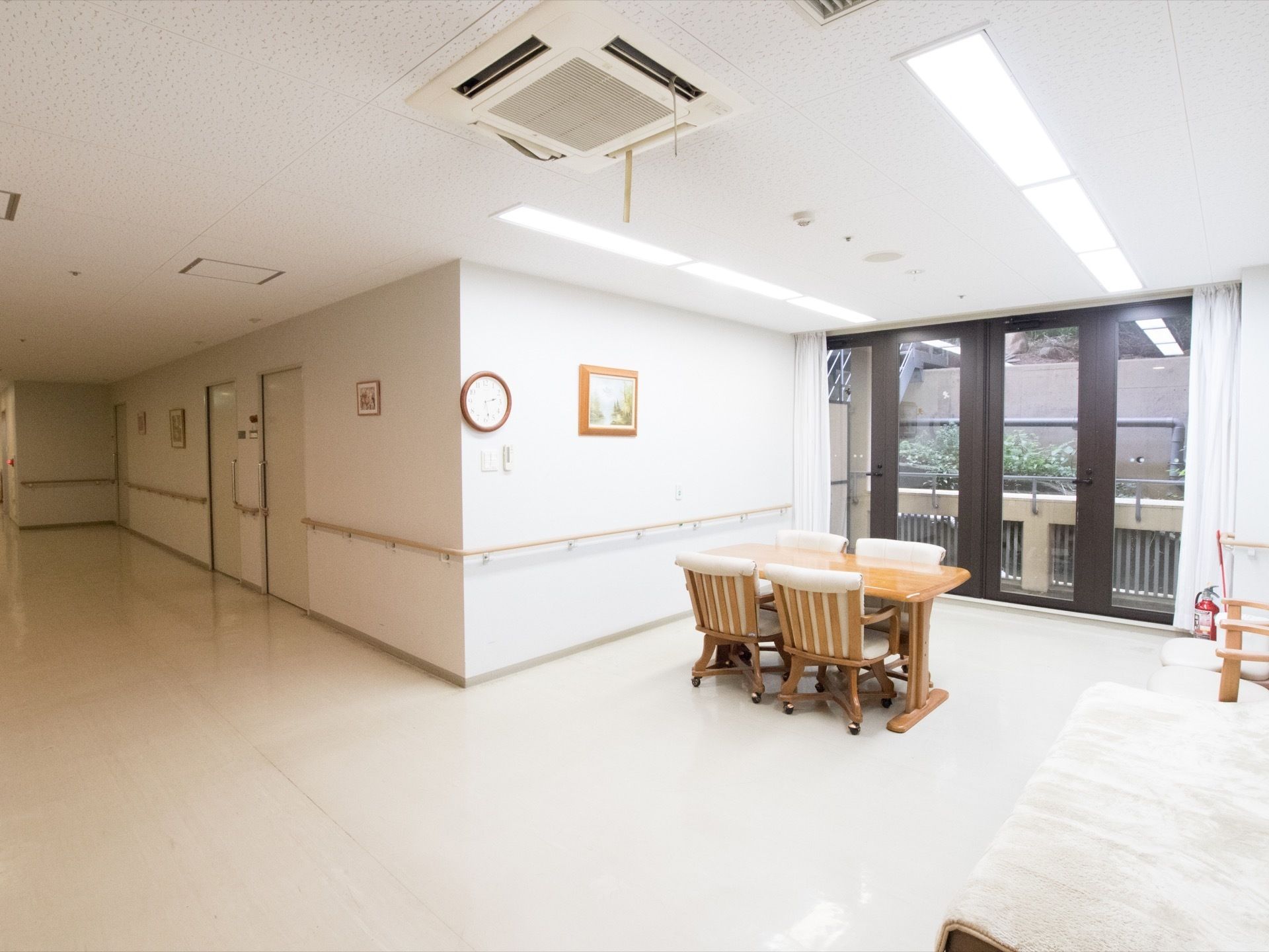ダイニングテーブルと椅子がセットで用意されている。大きな窓からはベランダが見えている。壁には時計と絵画が掛けられている。