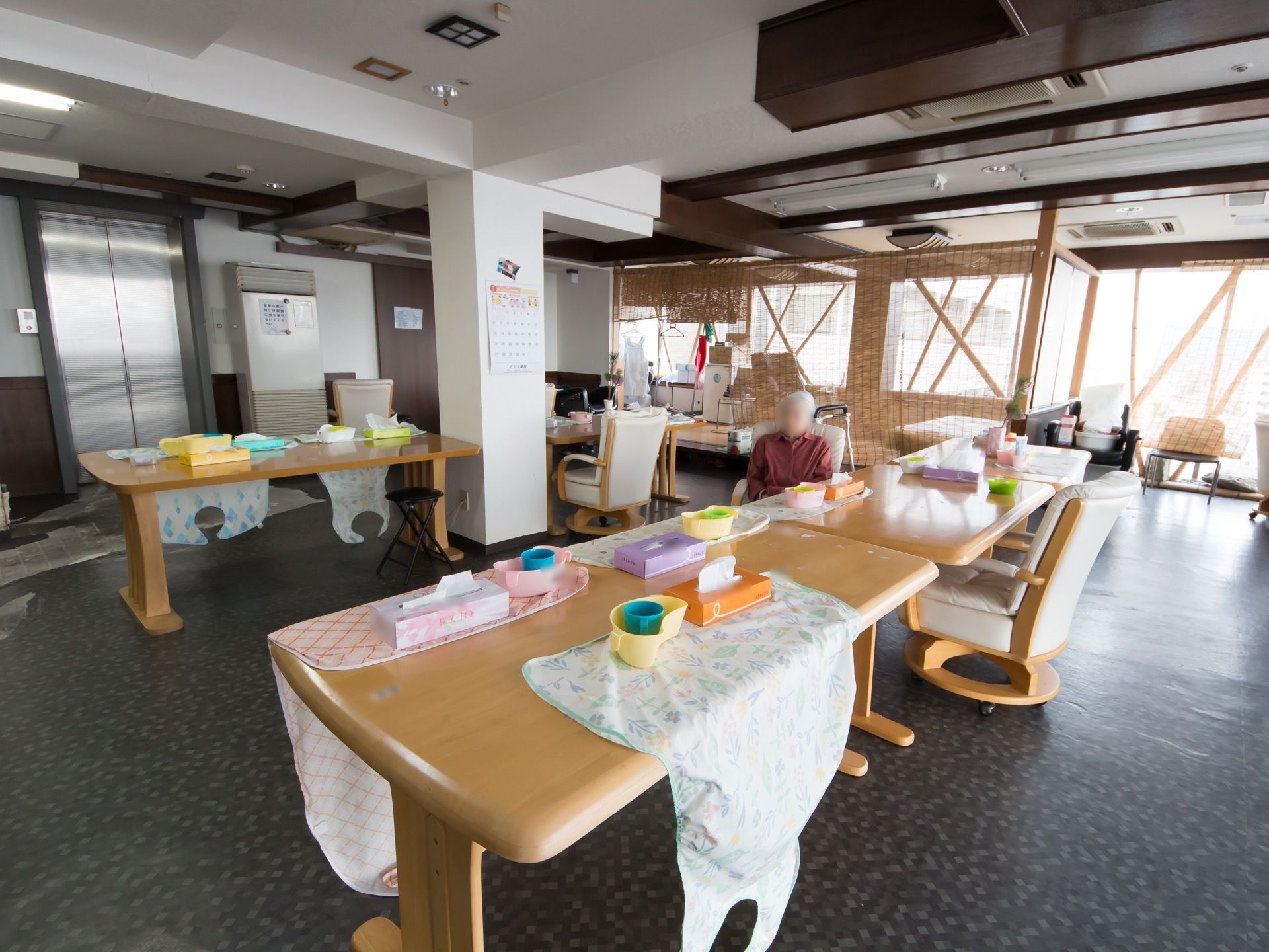 大きな窓が２面ある食堂。入居者様が食事の時に使用するエプロンの上に、食器やティッシュペーパーの箱が置かれている。