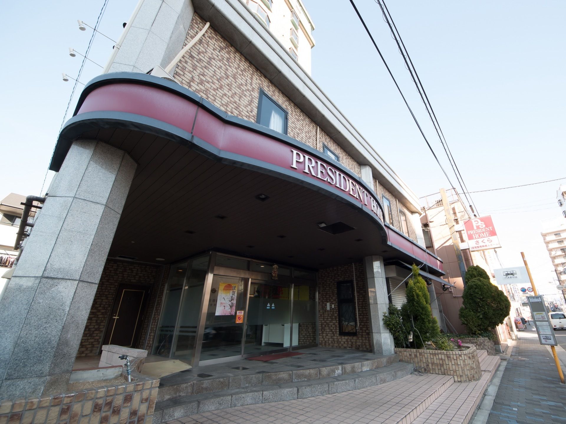 ホテルのエントランスのような玄関である。赤色の帯に施設名称が英語で表示されている。玄関の扉には時候の挨拶が掲示されている。