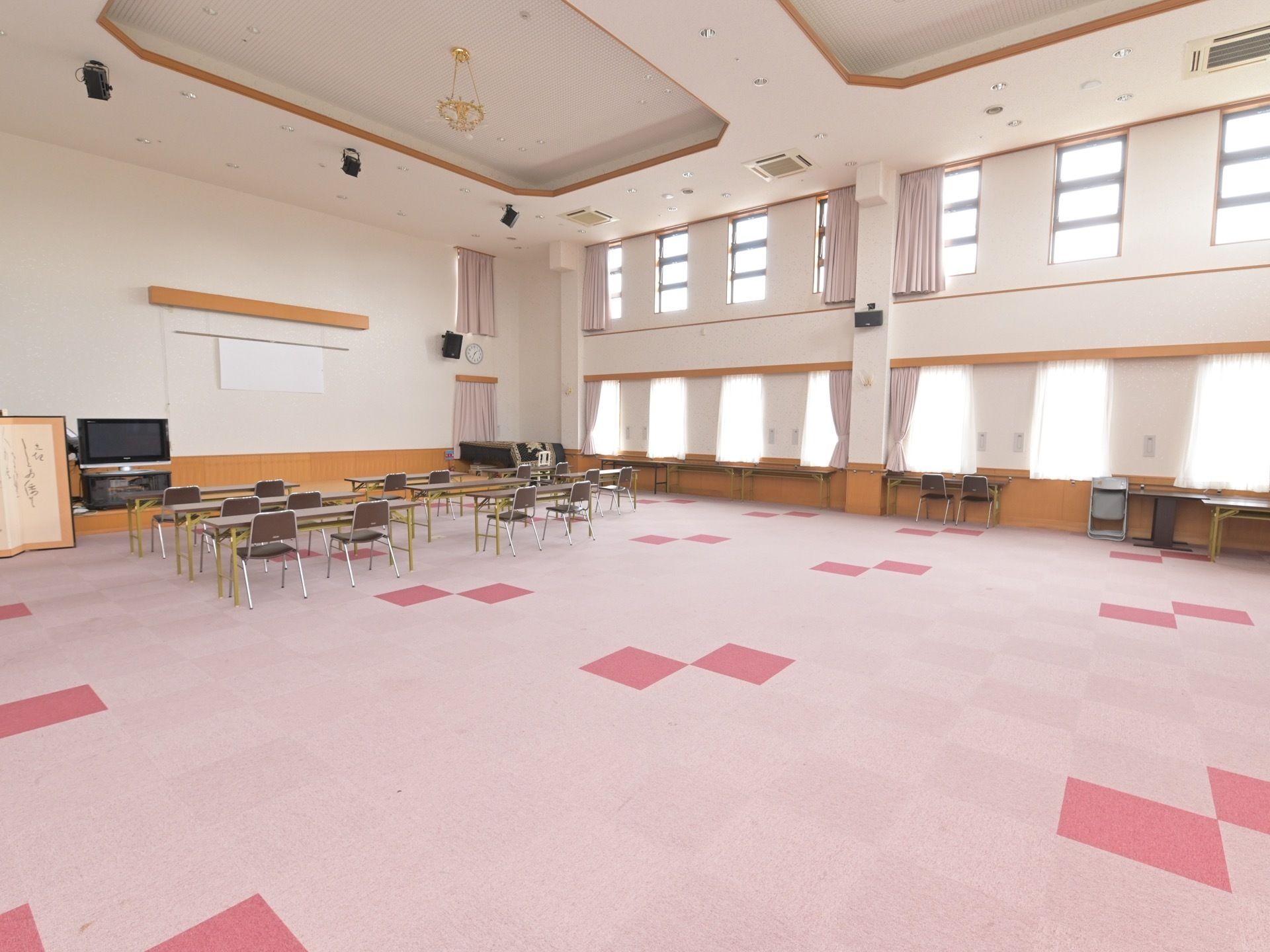 集会室の広い床面にはカーペットが敷き詰められている。前方に、複数の長机と椅子、液晶モニターが見える。