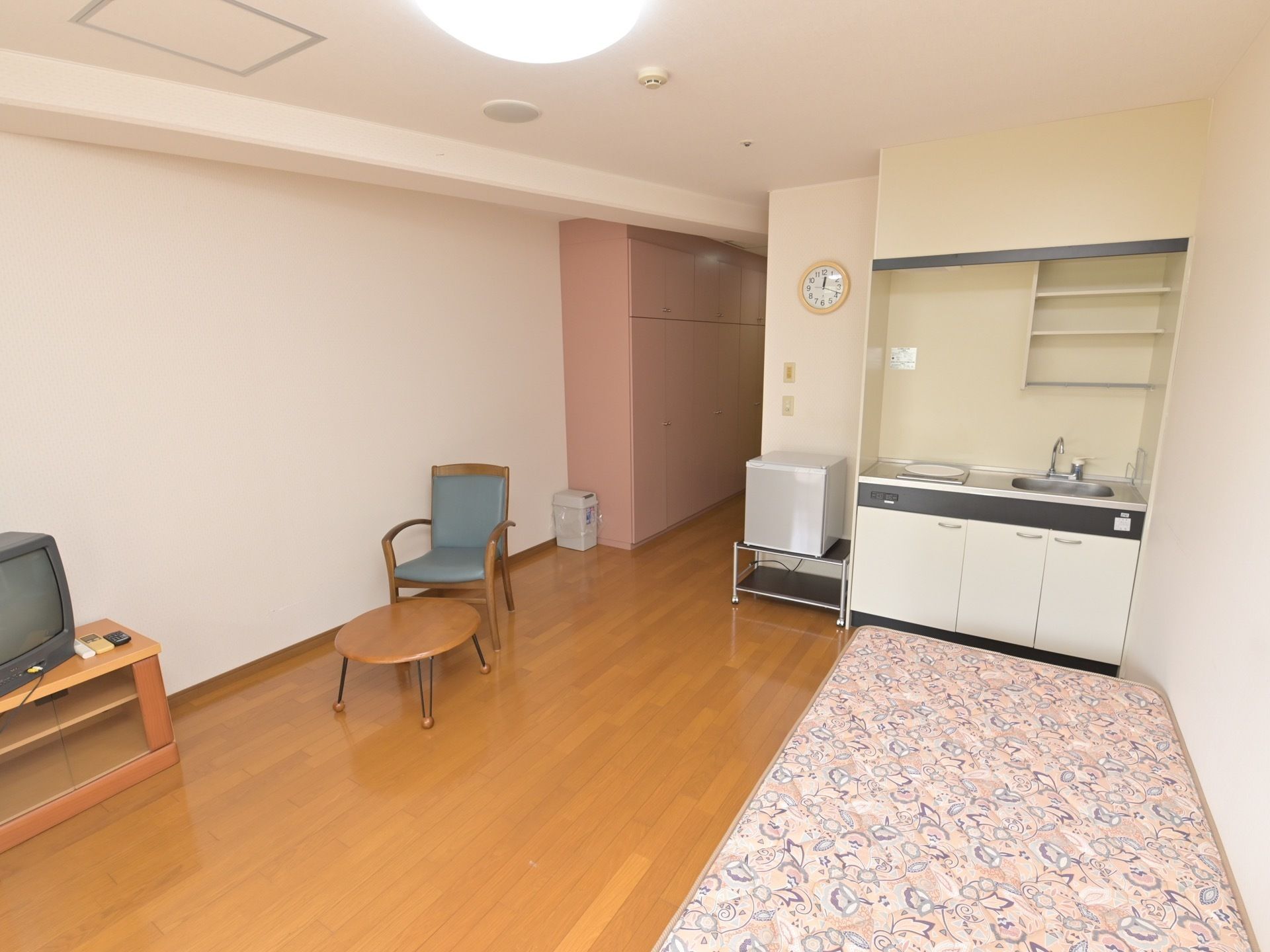 居室の通路に、天井に達する収納棚が設置されているのが見える。手前には、テレビやベッドなどが配置されている。