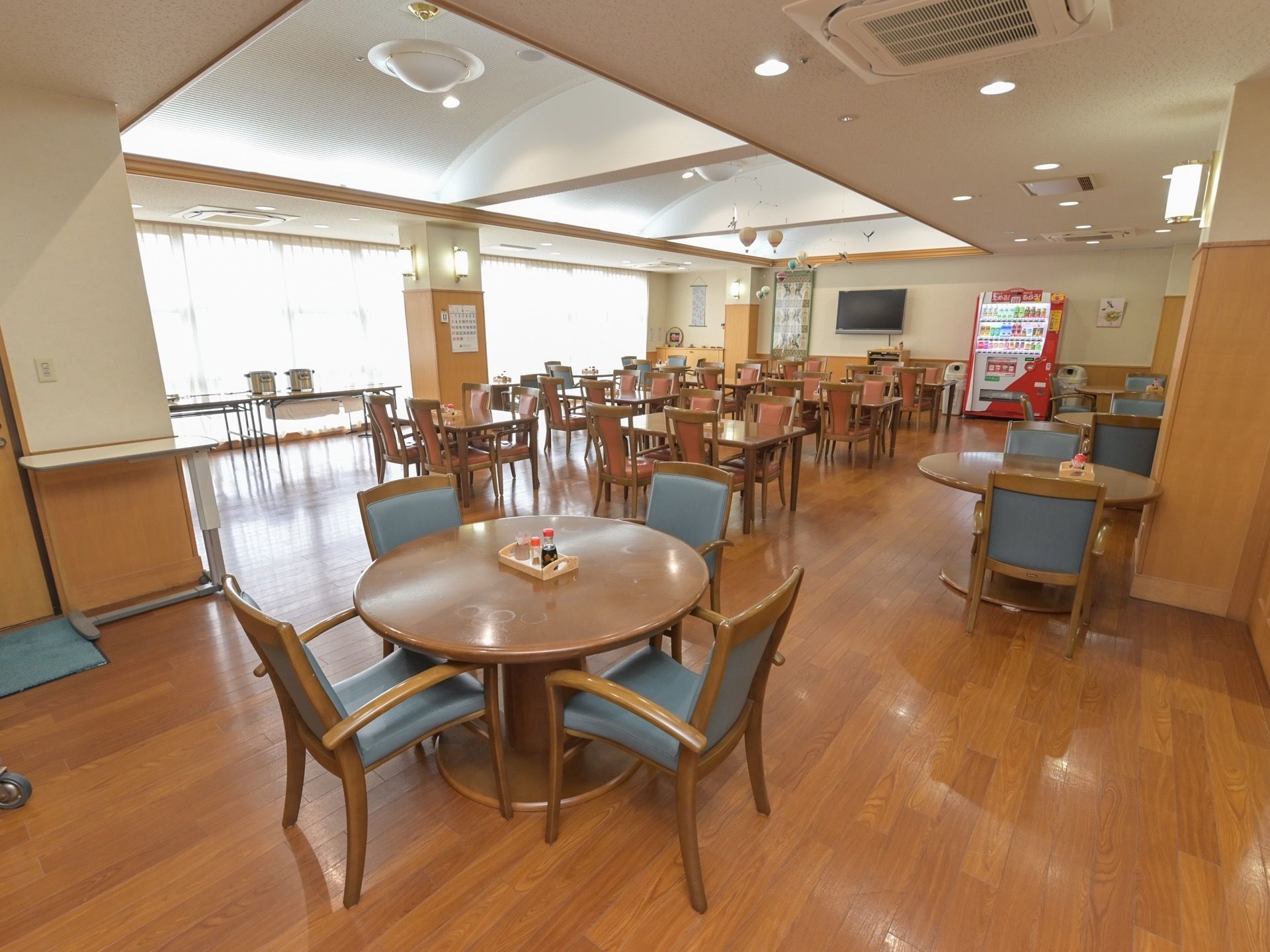 食堂の奧に、壁の高い位置に取りつけられた大型液晶テレビと飲料の自動販売機が見える。テーブルの天板は、四角いものと丸いものがある。