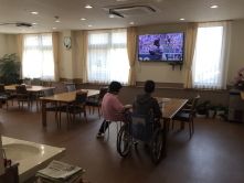 大きな壁掛けテレビがあり、車いすの高齢者が見ている。食堂にはダイニングテーブルが複数置かれている。