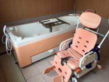 白とピンクの機械浴が置かれたタイル張りの浴室は明るい雰囲気。車いすタイプのリフトが置かれている。