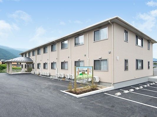 2階建ての建物はブラウン系でエル字型。広い駐車場が付き、側面には屋外階段も付いている。