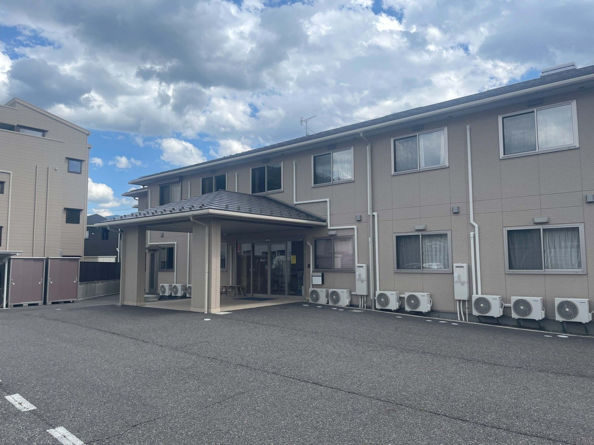 エル字型で広い駐車場が付いた2階建ての建物の玄関には屋根が設置されていて側面には屋外階段も付いている。