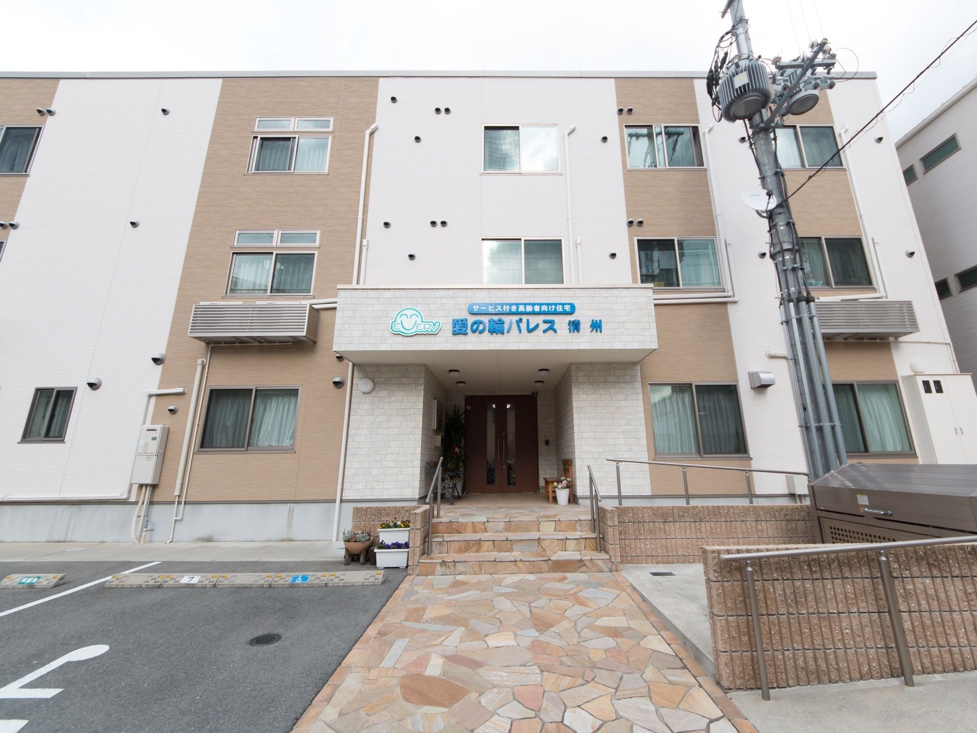 玄関前に設置された屋根部分に、青い文字で施設名が表示されている。外壁が２色になっており、落ち着いた雰囲気である。