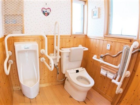 木目調の壁が優しい雰囲気のトイレ。男性用小便器と洋式トイレが並んで設置され、手すりも多くある。
