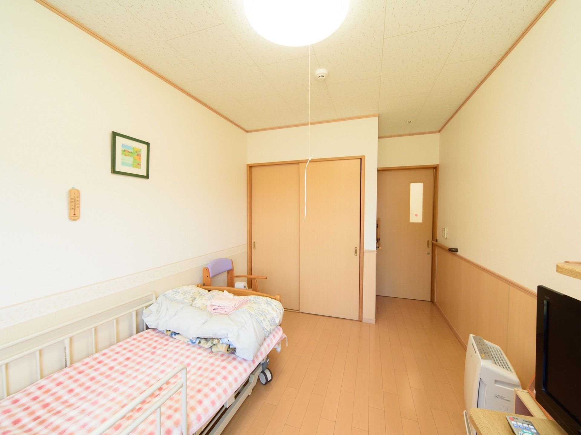 ベージュ色のフローリングと、同じ色合いの腰板でまとめられた明るい雰囲気の居室である。介護ベッドや押し入れが備わっている。
