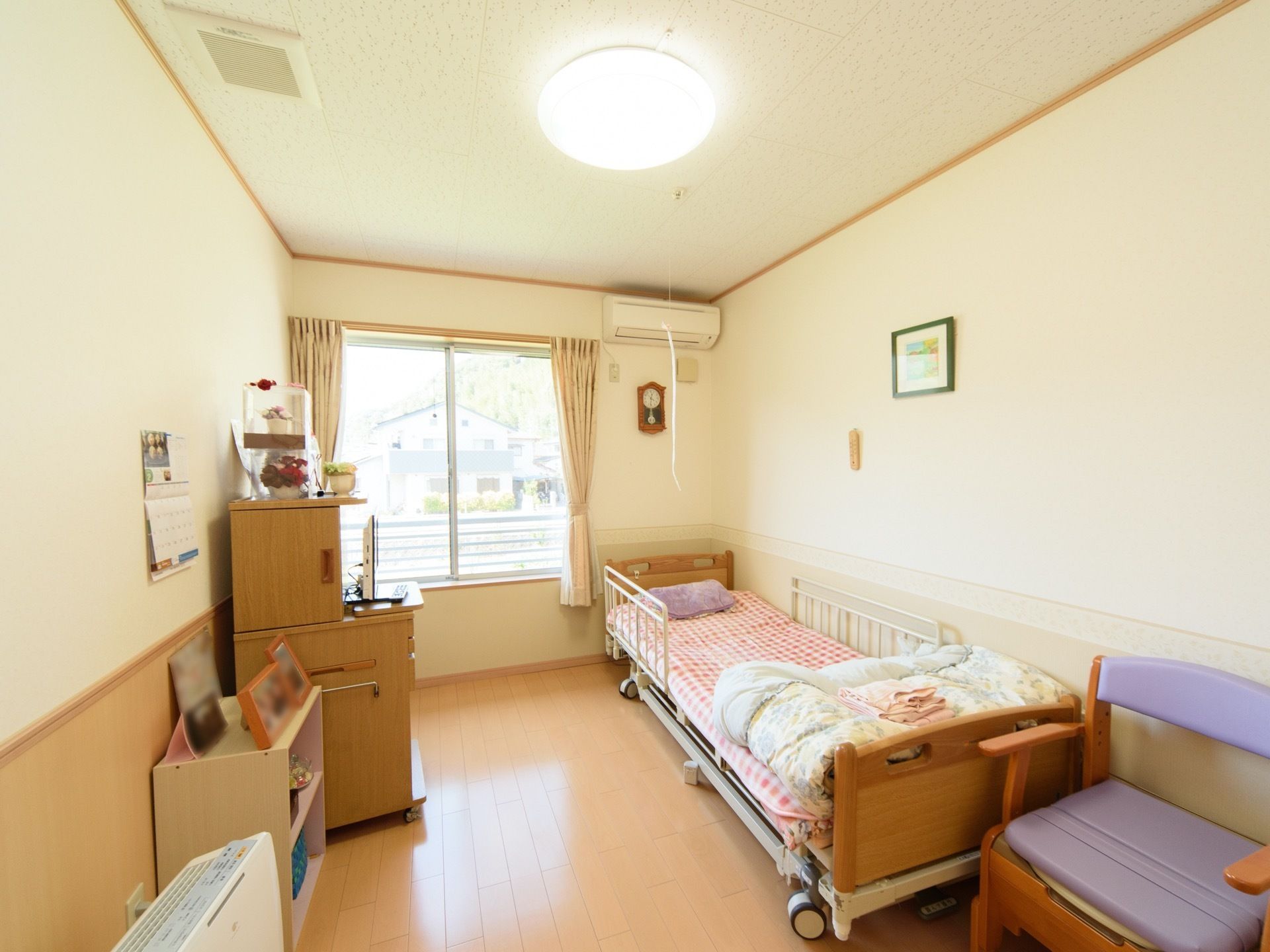 居室内に簡易式の椅子型トイレ、ベッド、収納棚などが置かれている。窓の近くにエアコンが設置されている。