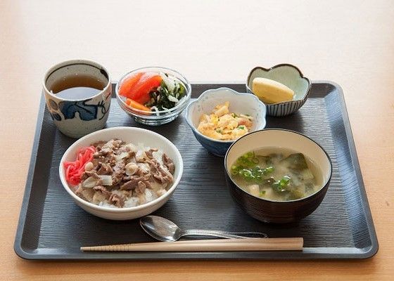 牛丼に味噌汁、サラダや煮物にフルーツとお茶が並んだ黒いトレーには、スプーンと箸が添えられている。
