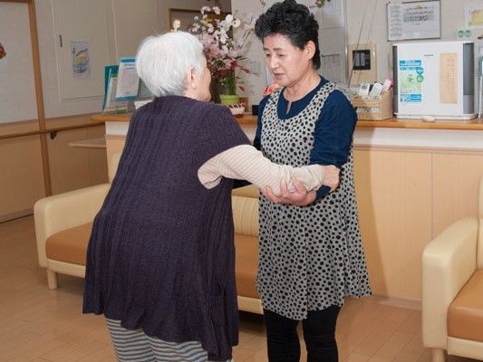 女性スタッフが女性入居者の腕を支え、歩行訓練を手伝っている様子。