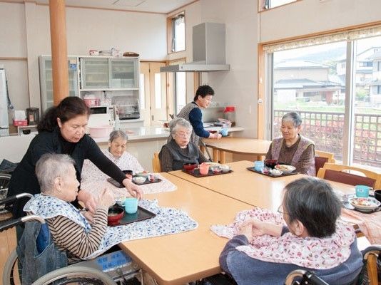 食事を楽しむ入居者の様子。料理が並べられたトレーを運んだり、配置を整えたりしているスタッフがいる。