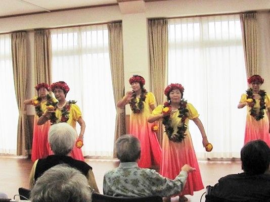 黄色のピンクのダンスに身を包んだ女性たちの踊りを見学する入居者の様子。