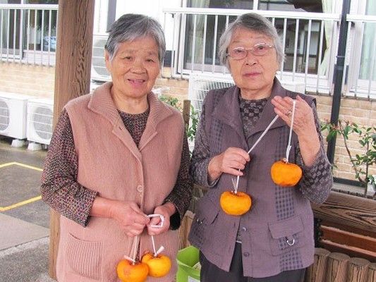 きれいに皮をむいてビニールひもを結び付けた柿を手にして微笑む入居者２名。