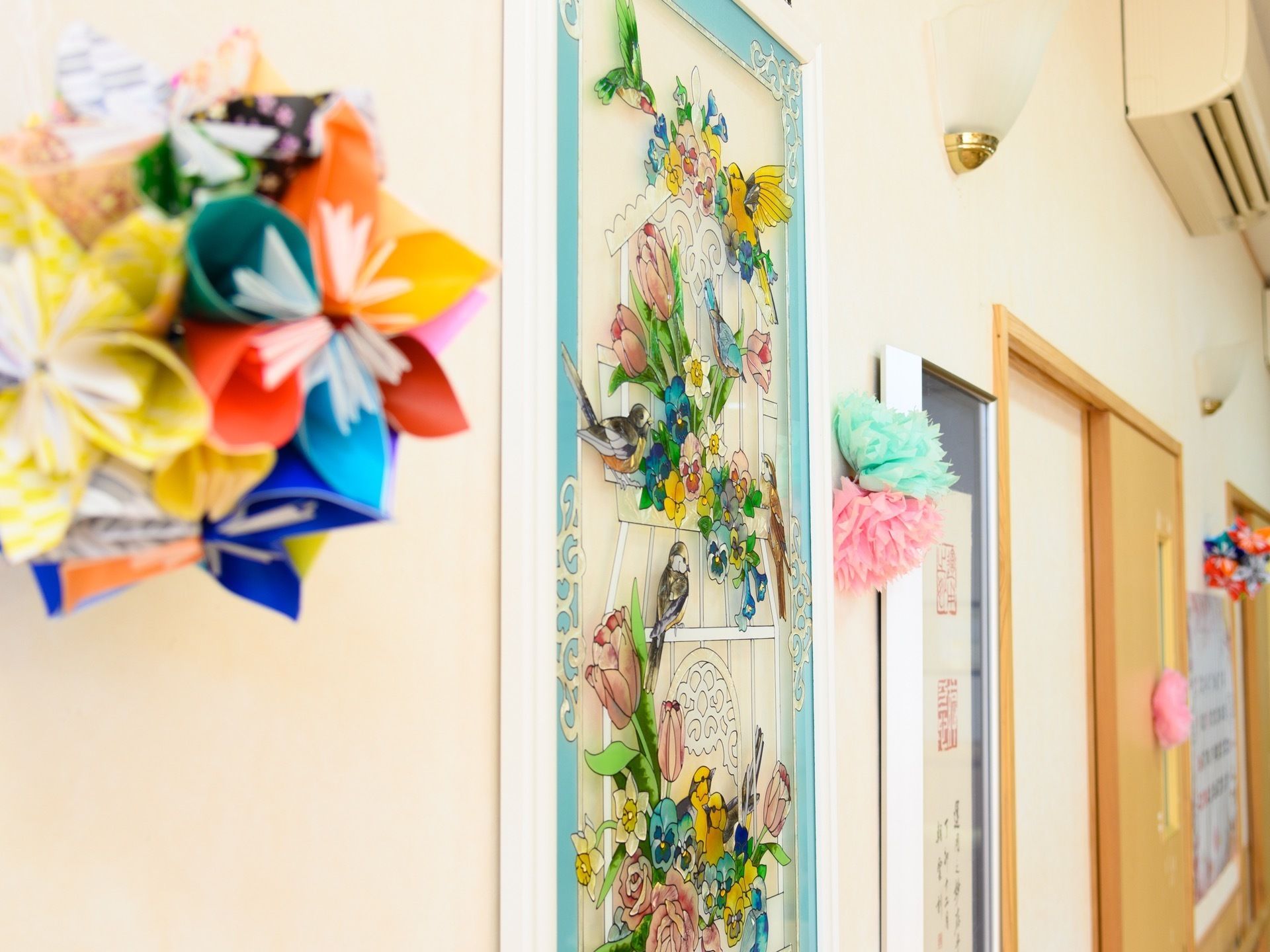 廊下の壁に、花や鳥をモチーフにした細工品やステンドグラス作品が飾られている。いずれも彩り豊かで、華やかな印象を与える。