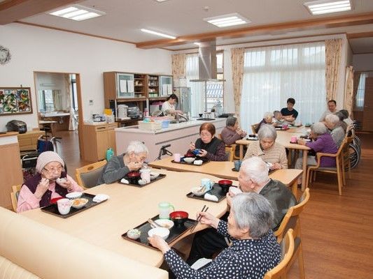 黒いトレーに並んだ料理を食べている入居者。スタッフは介助に当たったり、キッチンで作業をしていたりする。