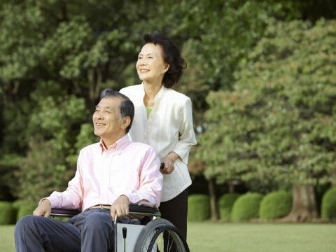 白いエプロンを付けた男性スタッフに付き添われて屋外で笑顔で過ごしている車いすの高齢女性。