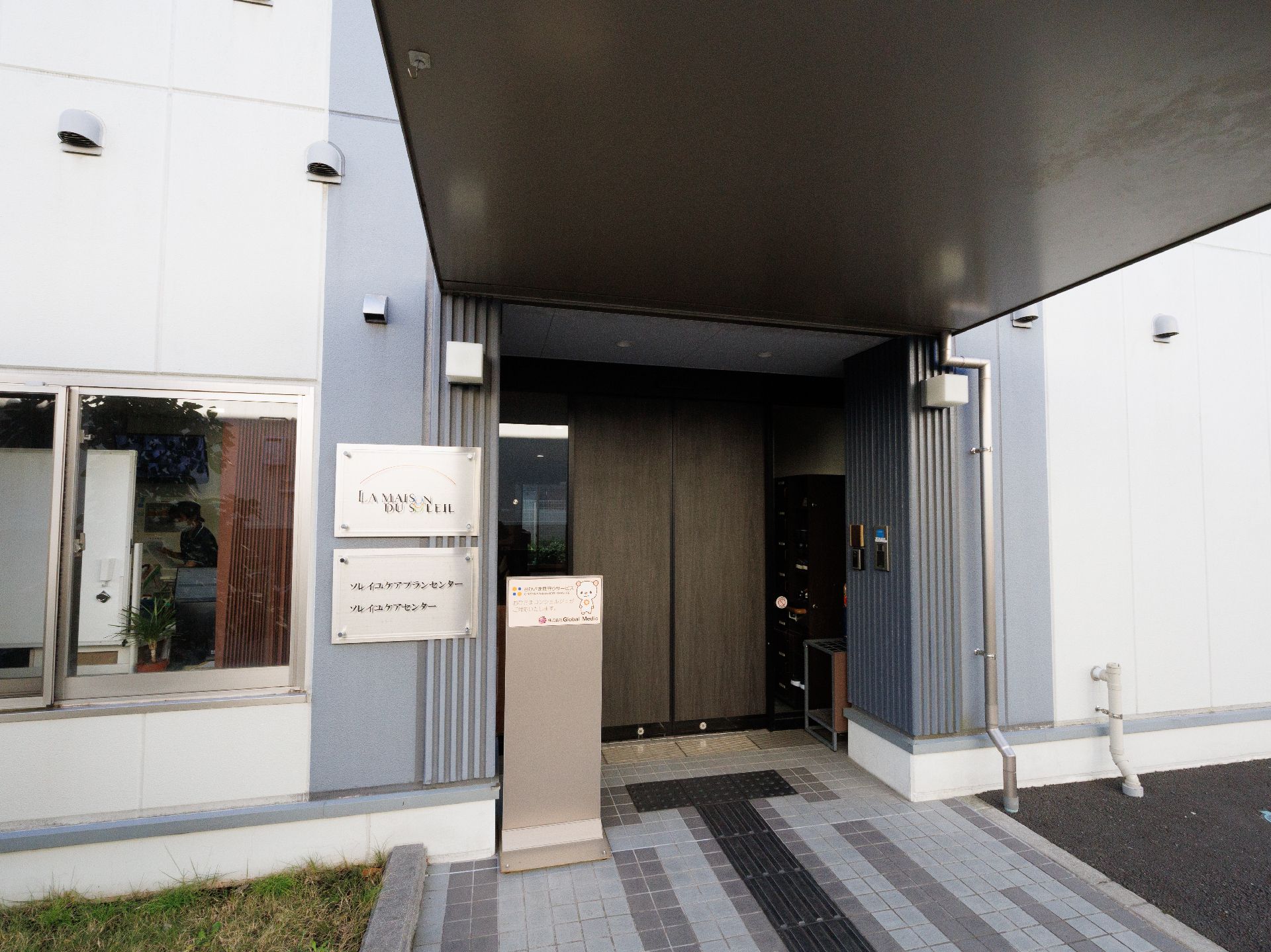 薬を飲もうとしている高齢女性のそばにはピンクのエプロンを付けた女性スタッフがいて、ほほえんでいる。