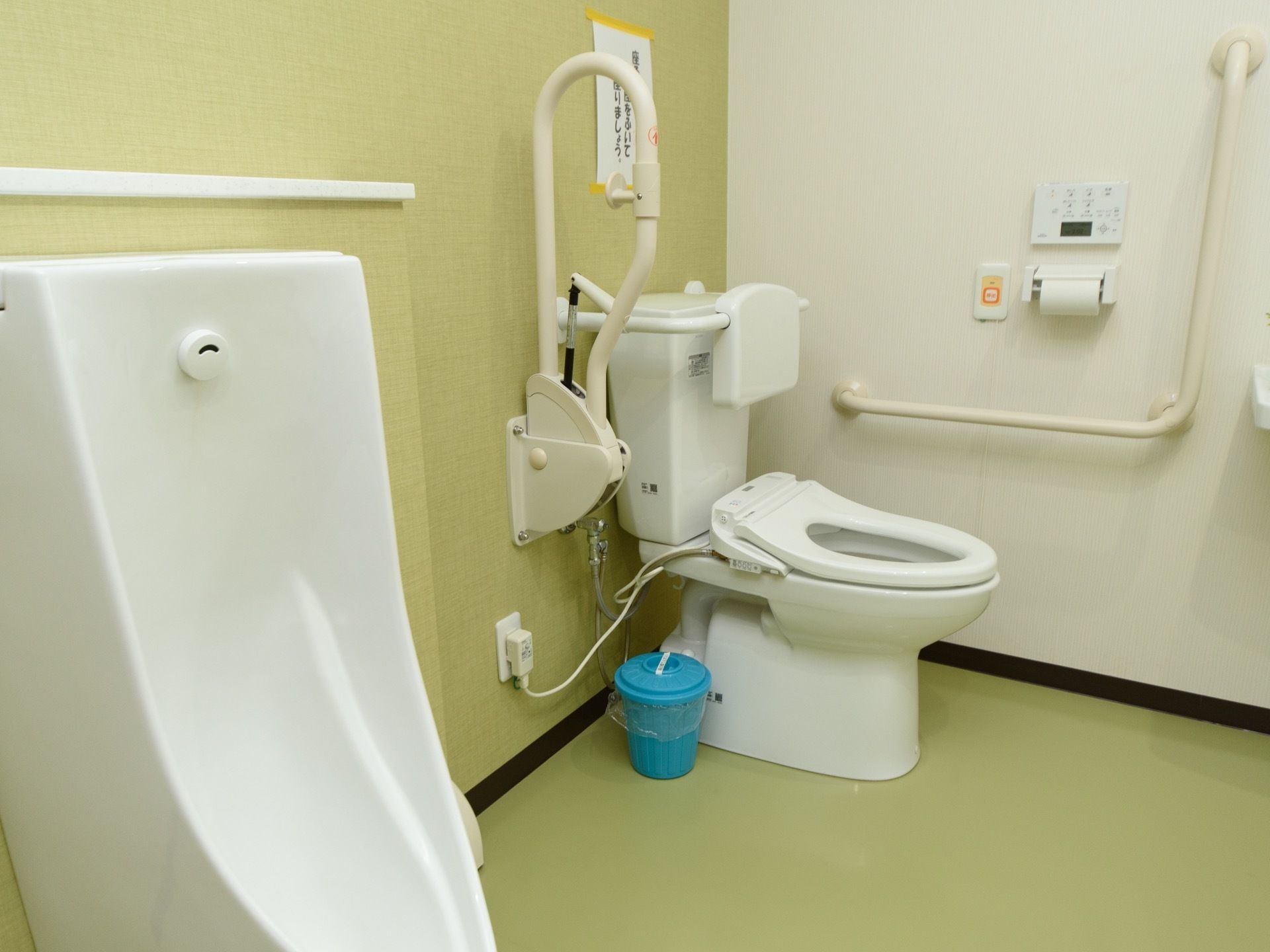 温水洗浄便座器が完備されている施設内のトイレである。可動式の手すりを間に、男性用の小便器が設置されている。