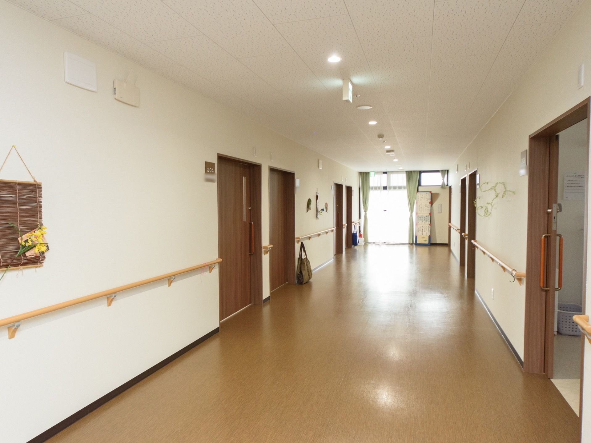 居室前の廊下は、幅広く設計されている。両サイドの白い壁には、ブラウン系の居室扉とネームプレート、手すりがある。