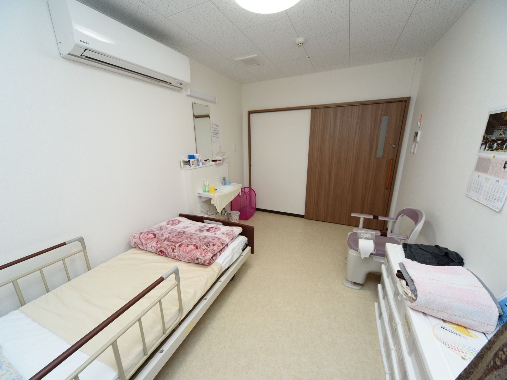 エアコンや洗面台が設置されている白い壁の居室である。介護用ベッドとポータブルトイレが配置されている。