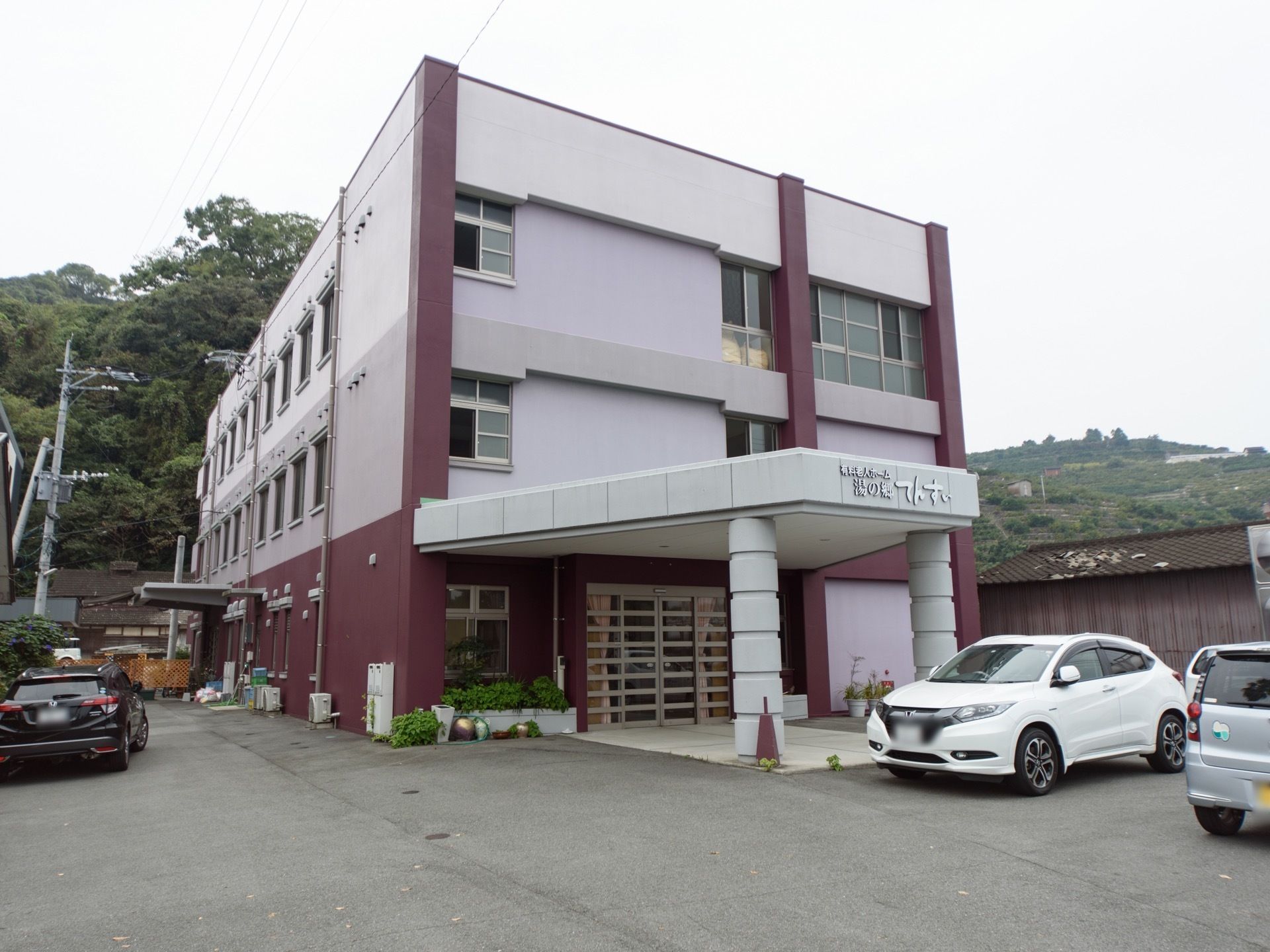 曇り空の日に山の緑を背景に立地する3階建の施設である。屋根部のある玄関ポーチが設けられ、前には白い乗用車が停車している。