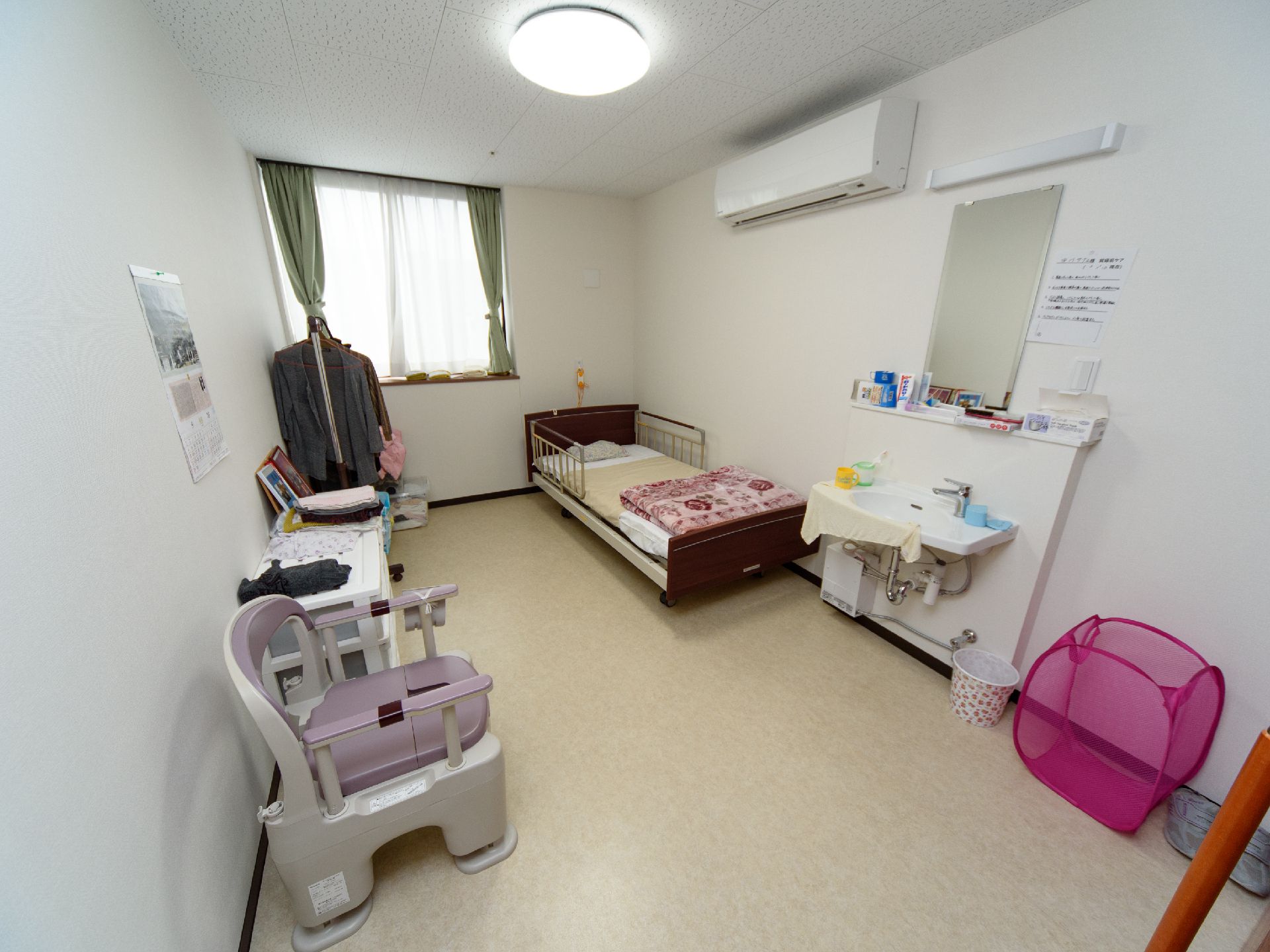 エアコンと洗面台が完備されており、壁際に介護ベッドやポータブルトイレが置かれている居室