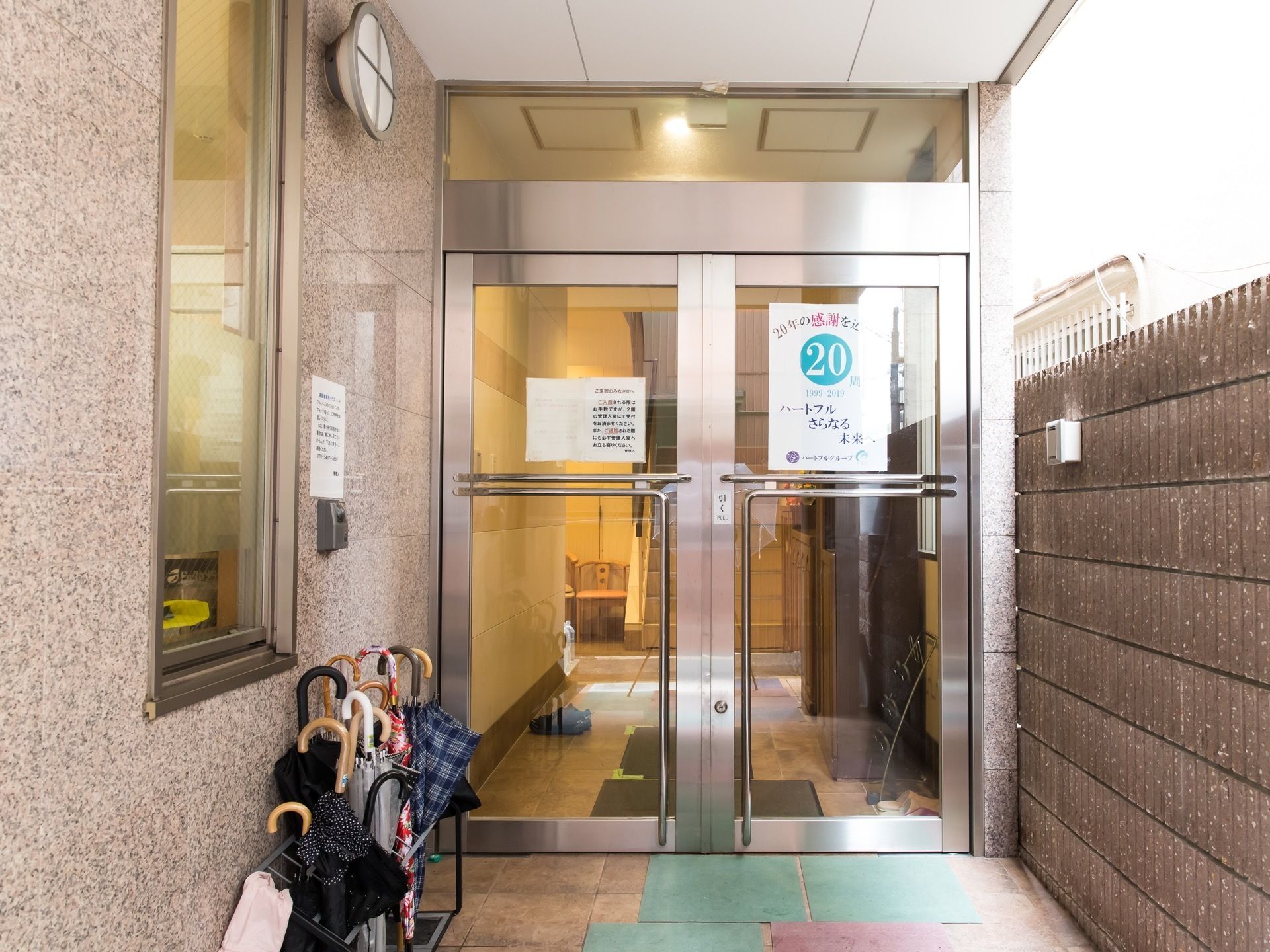 正面にガラス張りの開き戸があり、手前に傘立てが置かれている。建物の右側に接するように塀が設置されている。