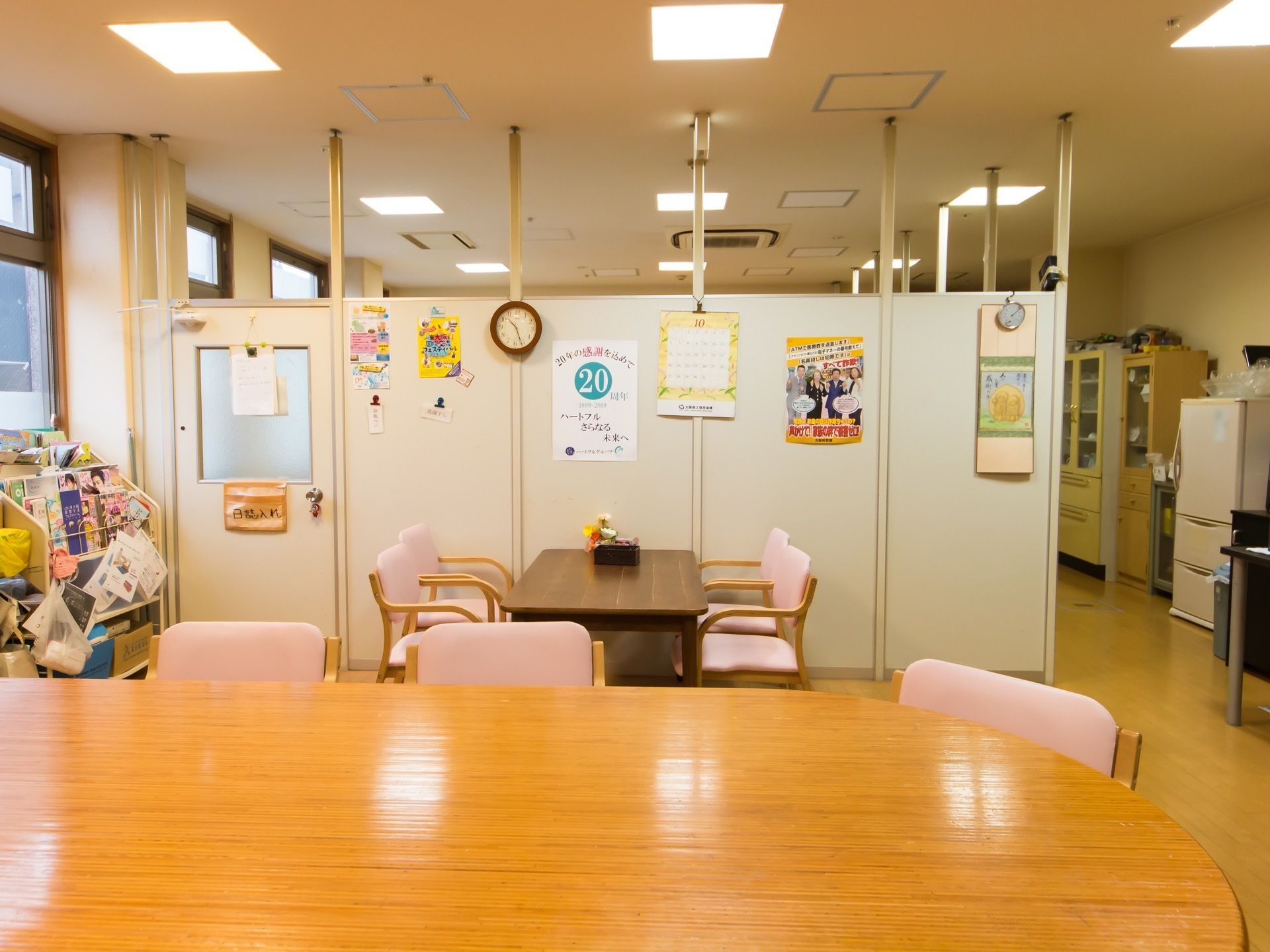 広間に木製のテーブルや介護用椅子が複数設置されている。右側に冷蔵庫や収納棚が置かれているのが見える。