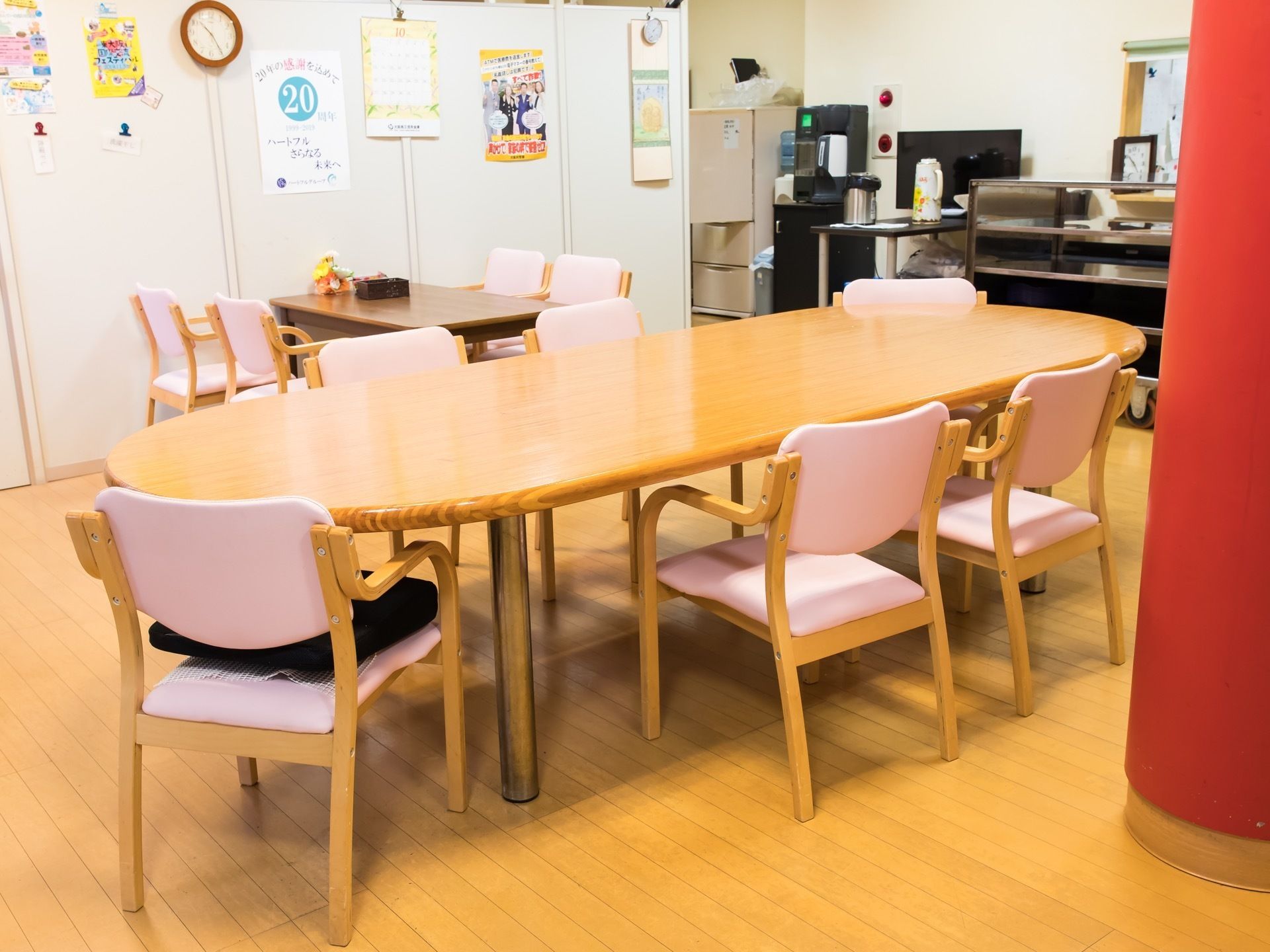 フローリングの広々としたスペースに、楕円形のテーブルと介護用椅子が設置されている。奥に冷蔵庫や配膳台が見える。