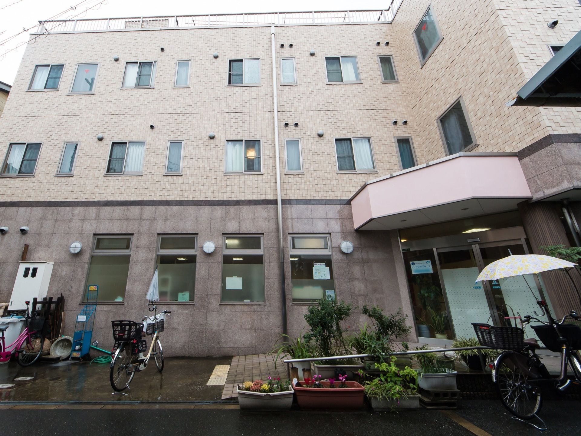 外壁に複数の窓が設置されている建物。玄関前のスロープの周囲には鉢植えが並んでおり、複数の自転車が停められている。