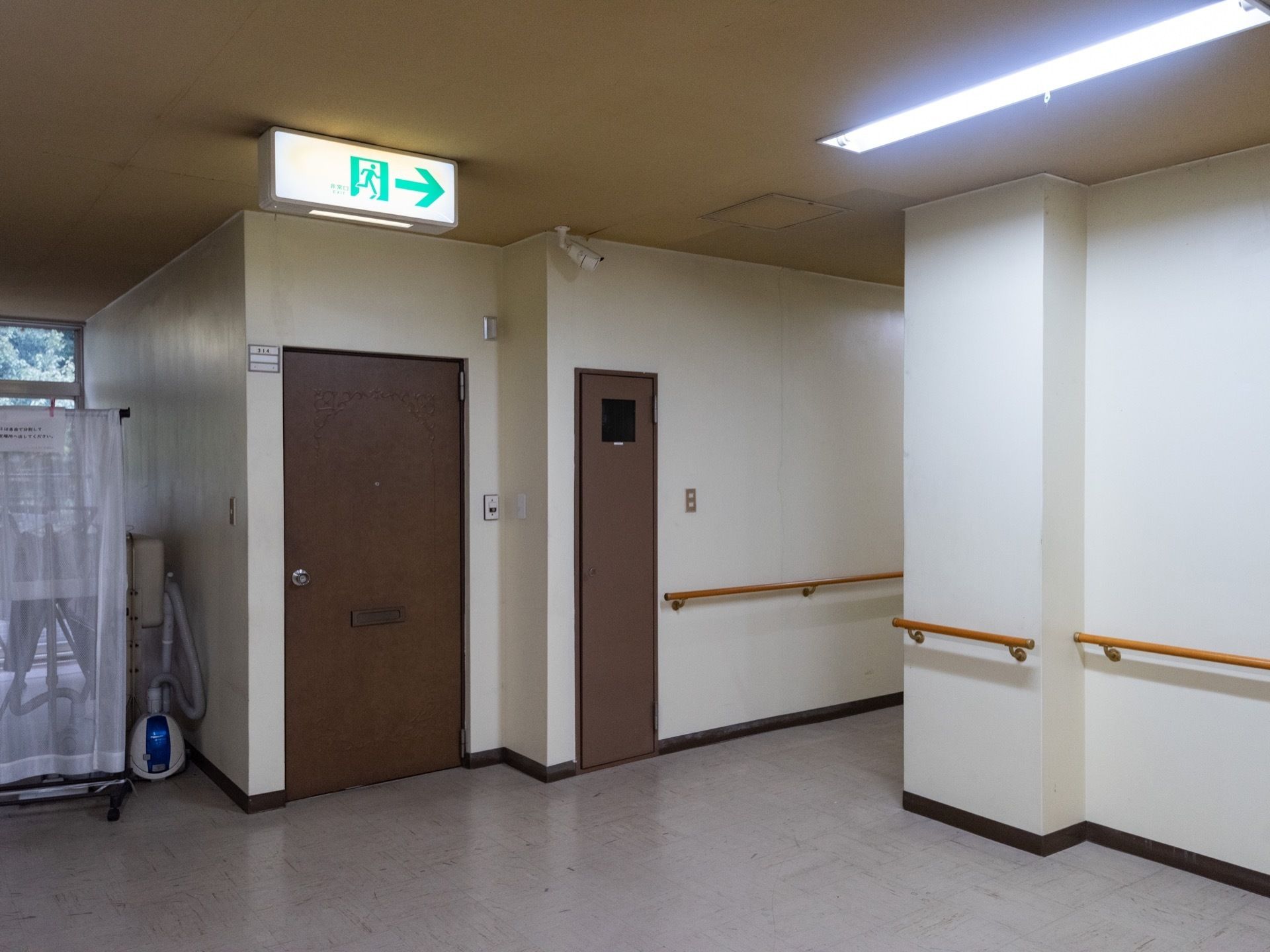 タイル貼りの廊下で、手前はホールになっており、右奥に続いている。正面にドアが２枚あり、天井に非常口の表示がされている。