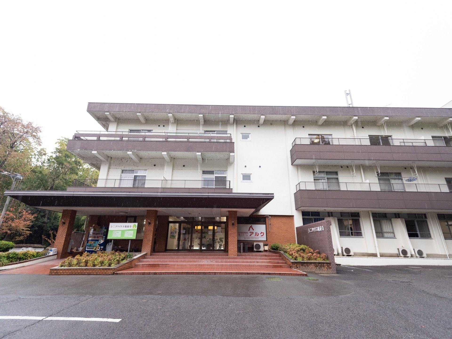 白い外壁にグレーの屋根がついた施設が奥に建っている。中央にレンガでできた玄関ポーチがあり、屋根が伸びている。