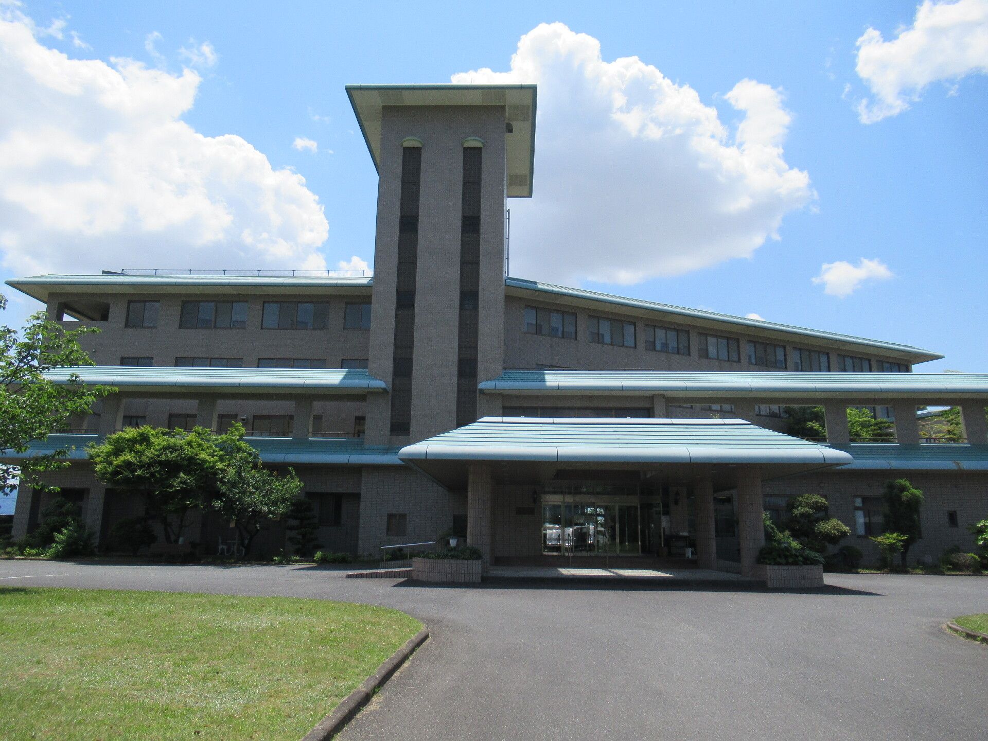 たくさんの大きな木に囲まれたブラウン系の外壁の建物には緑の屋根が付いている。