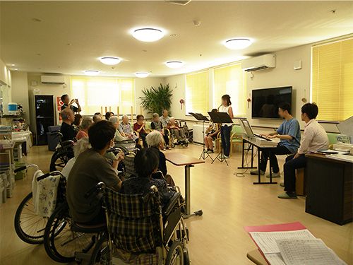 ボランティアによるコンサートの風景。電子ピアノなどを弾く演者が前にいて、それを囲むように入居者がいる。