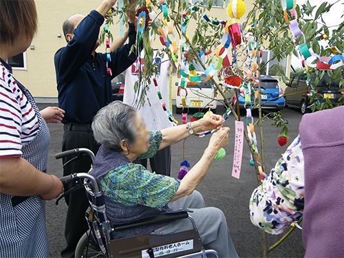 大きな笹に七夕飾りを取り付けようとしている女性入居者。車いすに乗っている。