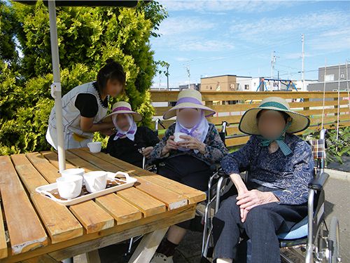 パラソルの下の木陰でティータイムを楽しむ３人の女性入居者と女性スタッフ。入居者は車いすに乗っている。